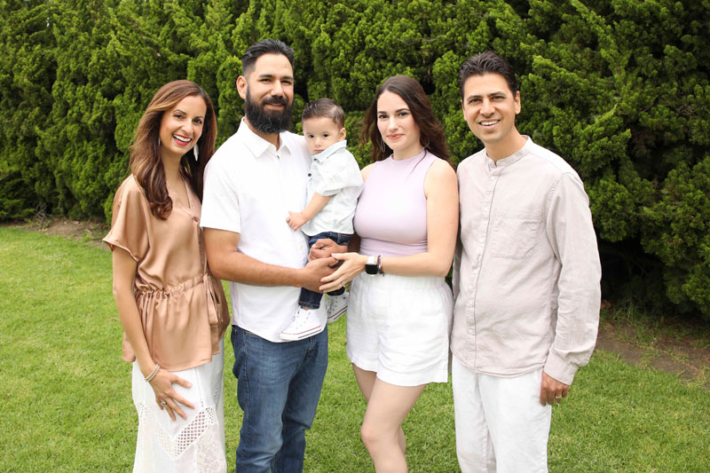 healinglight_tina_mukunda Five adults and a baby standing together on grass, smiling, with green trees in the background.
